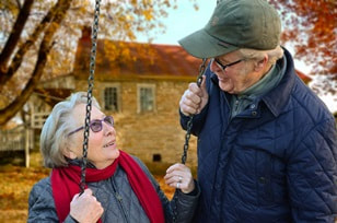 an older gentleman speaking to an older woman sitting on a swing.